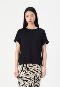 T-shirt noir à manches courtes avec une coupe décontractée, un col rond et des manches retroussées. Porté avec une jupe crème à motifs noirs.