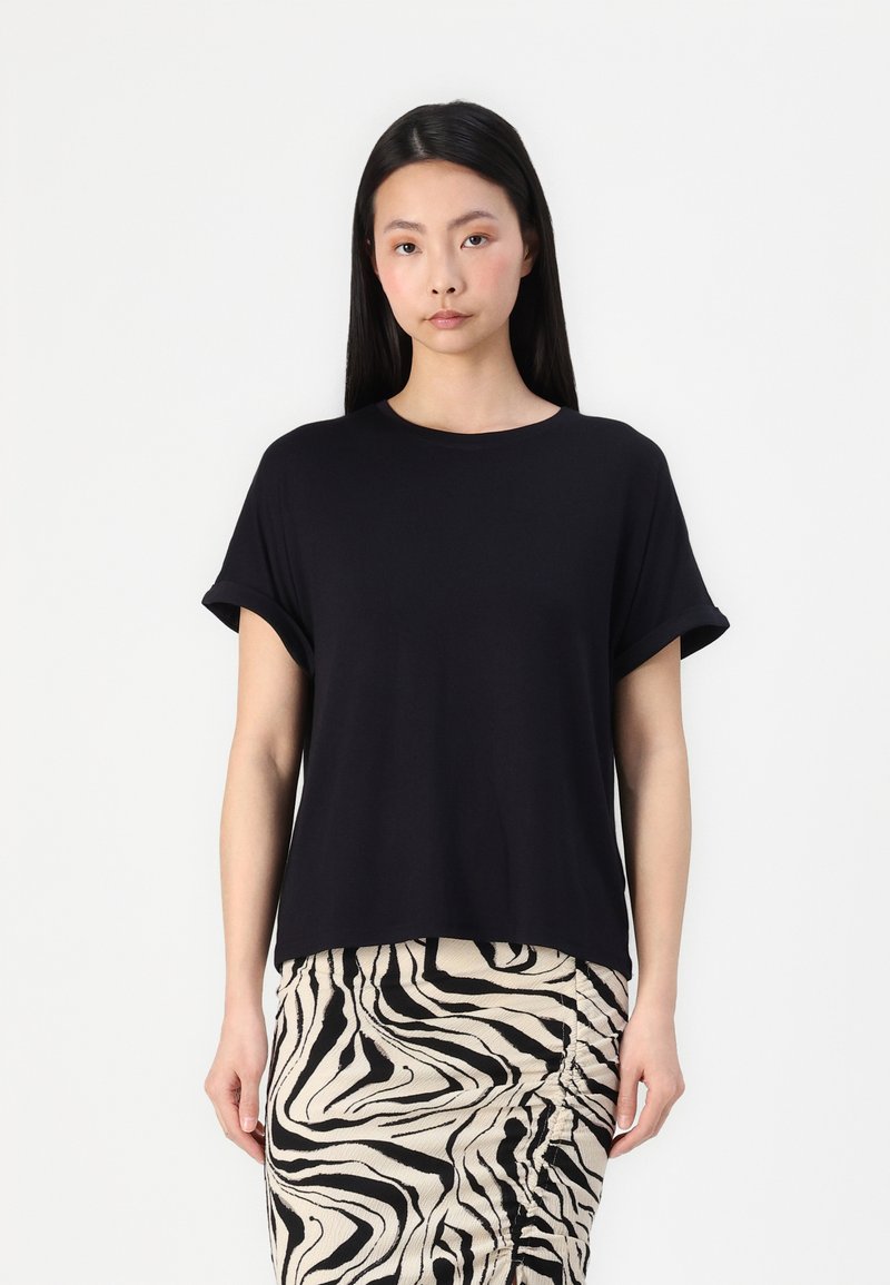 T-shirt noir à manches courtes avec une coupe décontractée, un col rond et des manches retroussées. Porté avec une jupe crème à motifs noirs.