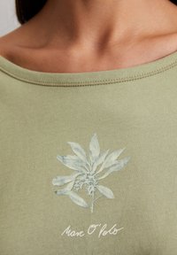 Olivgrünes Kurzarmoberteil mit einem hellblauen floralen Grafikdesign, das einen Stängel und Blätter zeigt, und weißem Text: "Marc O'Polo" darunter.