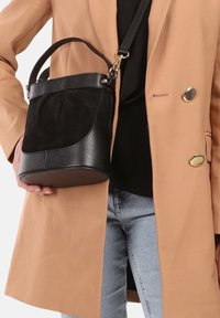 Sac seau en cuir noir et daim, avec un fond plat et un corps rond. Il dispose d'une sangle amovible et de détails en métal doré.