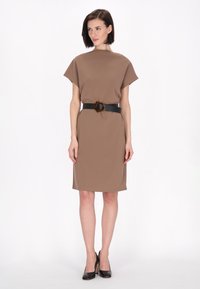 Ruskea mekko, jossa on korkea kaulus ja lyhyet hihat, vyötärölle kiinnitetty mustalla vyöllä, jossa on geometrinen solki.