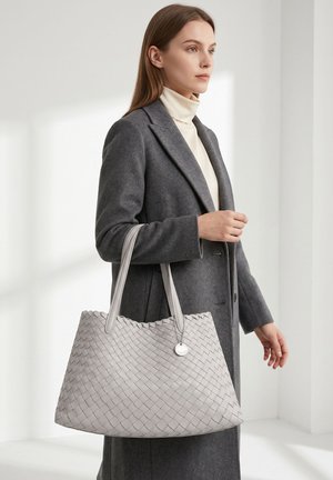 Femme portant un manteau gris et un col roulé crème, tenant un grand sac fourre-tout tissé gris clair à double poignée à l'intérieur.