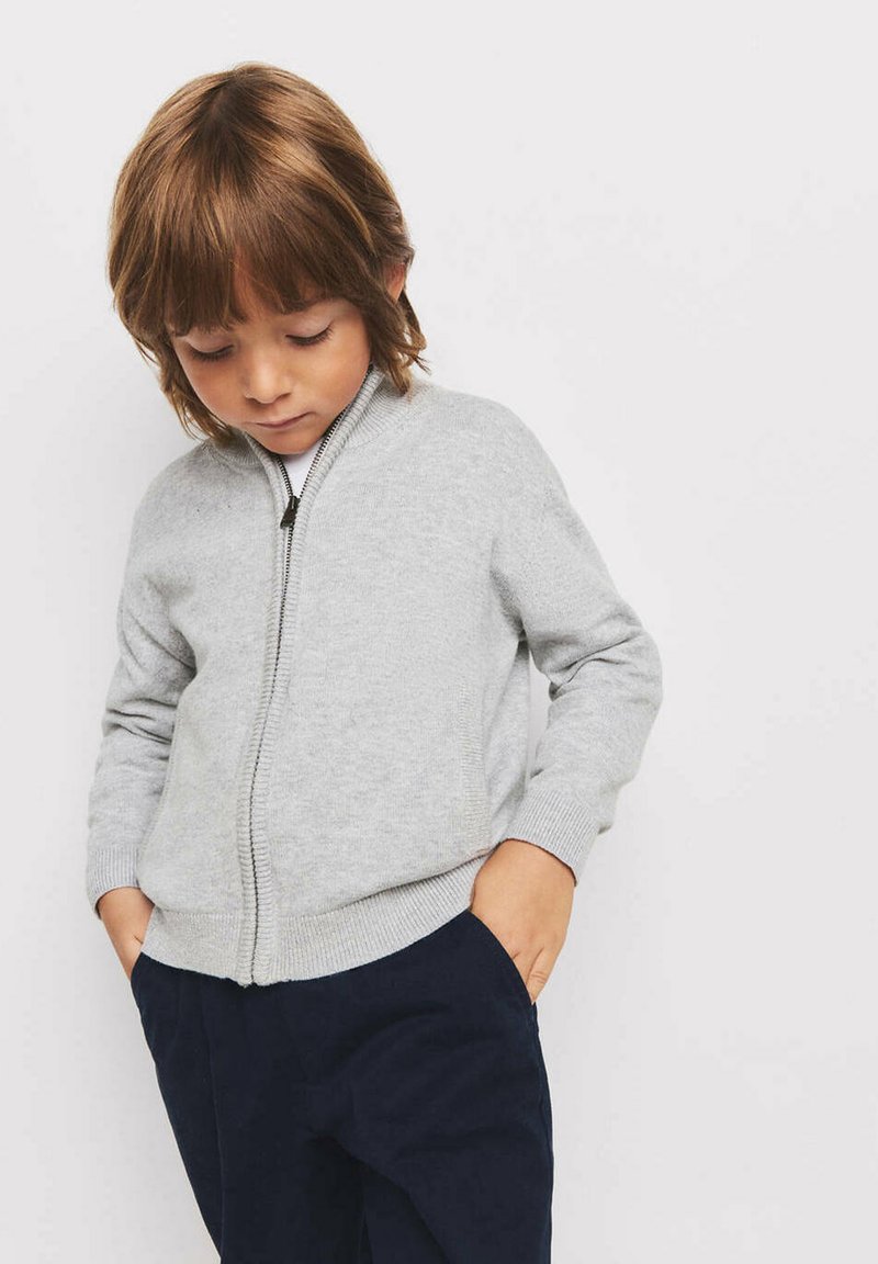 Jeune enfant aux cheveux châtain clair portant un sweat gris zippé et un pantalon foncé, les mains dans les poches, regardant vers le bas.