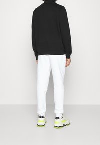 Persona vestida con una camiseta de manga larga negra, pantalones blancos y zapatillas blancas con detalles en amarillo neón, de pie frente a una pared blanca lisa.