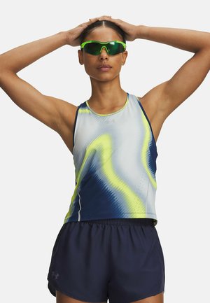 Vrouw met groene sportzonnebril, een mouwloos sporttopje met blauwe en gele patronen en donkerblauwe korte broek, handen rustend op haar hoofd.