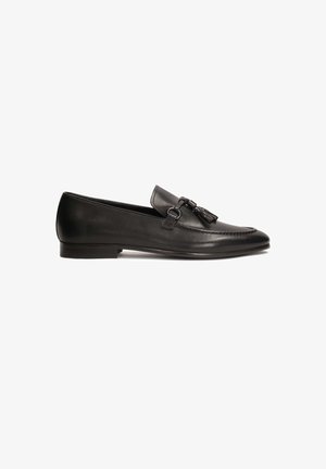 Des mocassins en cuir noir avec un talon plat, présentant un détail de gland décoratif et une boucle à l'avant, texture lisse et forme épurée.