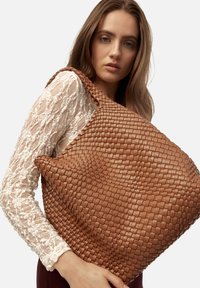Sac fourre-tout en cuir tissé de couleur marron chaud, présentant une surface texturée et une forme large et plate. Conçu avec une bandoulière robuste.