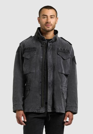 Young man wearing a faded black military-style jacket with multiple front pockets and epaulettes, standing against a plain light background.