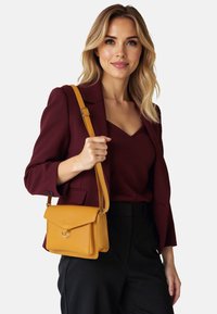 Vrouw draagt een bordeauxblazer en top, zwarte broek, houdt een mosterdgele schoudertas met verstelbare band vast.