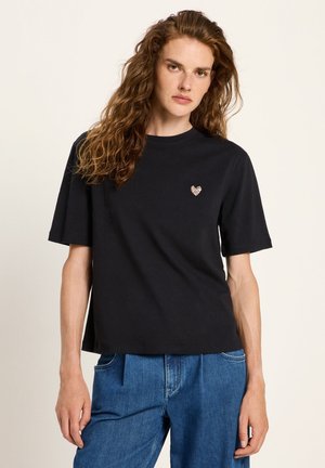 Woman with long curly hair wearing a black short-sleeve shirt with a small heart emblem and blue jeans against a plain background.