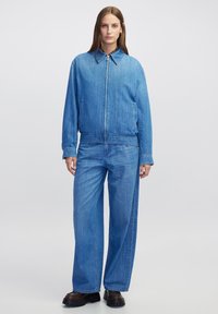 Vrouw die staat en een blauwe denim jack met rits en bijpassende wijdlopende spijkerbroeken draagt, met zwarte chunky schoenen, tegen een eenvoudige achtergrond.