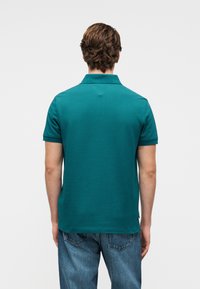 Τιρκουάζ πόλο μπλουζάκι με κοντά μανίκια, γιακά και λεία υφή. Διαθέτει άνετη εφαρμογή και συνδυάζεται με μπλε τζιν. Εμφανίζεται η όψη από πίσω.