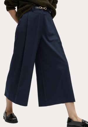 Pantalones cropped de pierna ancha en azul marino combinados con mocasines de charol negro y un suéter texturizado verde oscuro.