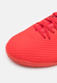 Červený sportovní bot, který má texturovaný syntetický svršek, zaoblenou špičku a kontrastní tkaničky. Podešev má mírně opotřebovaný okraj.