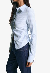 Lichtblauw gestreept shirt met een op maat gemaakte kraag, knopen aan de voorkant en geplooide details aan de zijkanten. G gedragen met donkere jeans.