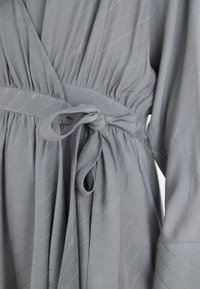 Robe portefeuille grise en tissu texturé avec des lignes diagonales, une taille élastique et un détail de ceinture nouée. Manches longues avec un drapé doux.