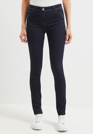 Kvinde iført mørkeblå skinny jeans, hvide sneakers og en hvid top, stående med armene afslappet ned langs siden.