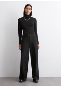 Schwarzes Rollkragenoberteil, figurbetontes Design. Schwarze, weit geschnittene Hosen mit vertikalen Streifen, hohe Taille und Gürtelschlaufen. Schwarze Schuhe. Neutraler Hintergrund.