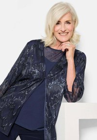 Dunkelblaue, leichte Jacke mit durchsichtigen Ärmeln und Paillettenverzierungen, die über einem einfarbigen navyblauen Oberteil getragen wird und ein strukturiertes Erscheinungsbild bietet.