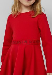 Czerwona sukienka z długim rękawem i rozkloszowanym dołem. Posiada teksturowany pas z błyszczącym haftem z napisem "S. OLIVER". Miękka tkanina.