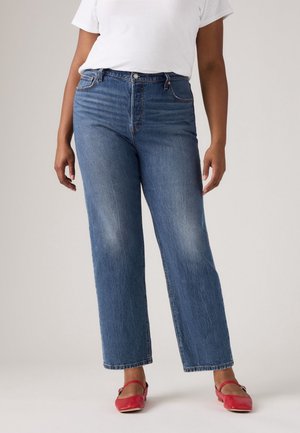Frau trägt hochtaillierte blaue Jeans mit geraden Beinen, ein weißes T-Shirt und rote flache Schuhe und steht vor einem neutralen Hintergrund.