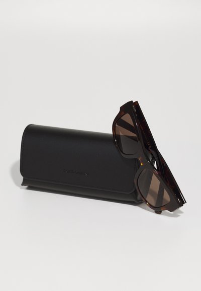 Brown tortoiseshell sunglasses with dark lenses sit next to a black leather case, featuring embossed branding on the surface.