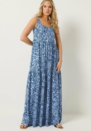 Kobieta o długich blond włosach, ubrana w bezrękawną, niebiesko-białą sukienkę maxi z wzorem i sandały, stojąca na tle jednolitego jasnego tła.