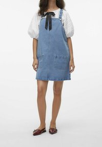 Kvinna som bär en blå denimpincettklänning över en vit blus med puffärmar och en svart rosett, i kombination med bordeauxfärgade spetsiga ballerinas.