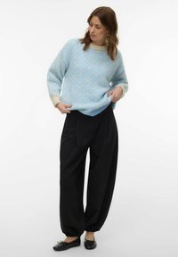Femme portant un pull bleu clair à motifs avec des poignets et un col crème, un pantalon noir ample et des ballerines noires sur fond blanc.