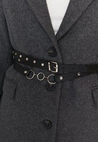 Zwarte leren riem met metalen ronde schakels, gedragen over een grijze wolmix jas met zwarte knopen en een gestructureerde afwerking.