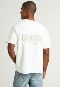 Baldas baltas medvilnės marškinėliai, ant nugaros su šviesiai pilkos spalvos "CHASIN'" grafika. Trumpomis rankovėmis, laisvas kirpimas, apvalaus kaklo dizainas. Dėvimi su mėlynomis džinsinėmis kelnėmis.