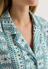 Turquoise bedrukte pyjamato met een kraaglijn. Bevat verschillende geometrische en bloemmotieven op een lichte achtergrond. Zachte katoenen textuur.