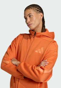 Giacca arancione con cappuccio realizzata in tessuto morbido, dotata di una zip frontale, strisce a contrasto sulle maniche e un logo sul petto.