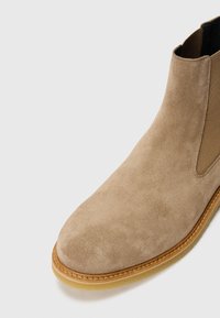 Botte ankle en suède beige avec des panneaux élastiques sur les côtés et un bout rond. Dispose d'une semelle légère avec un bord cousu pour plus de durabilité.