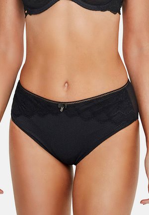 Femme portant des sous-vêtements noirs à bordure en dentelle et un soutien-gorge assorti, montrant le ventre et le haut des cuisses sur un fond uni.