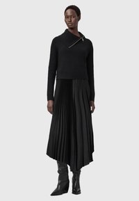 Μαύρος πλεκτός πουλόβερ με πλατύ γιακά και ασημί φερμουάρ, συνδυασμένος με πτυχωτή μαύρη φούστα και μαύρες ψηλές μπότες με τακούνι.