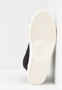 Chaussure noire inclinée vers le haut, montrant une semelle blanche avec des motifs de gomme circulaires et linéaires sur fond blanc.