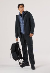 Black waterproof jacket over a gray fleece top and black pants, paired with black shoes. A black backpack is held in one hand.
