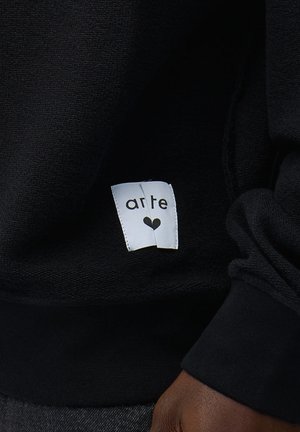 Tessuto nero con un'etichetta bianca su cui è scritto "arte" sopra un piccolo cuore nero, indossato su un indumento a manica lunga con polsino e manica visibili.