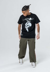 T-shirt nera con grafica di teschio e design di pugnale, abbinata a pantaloni cargo verde oliva. Sneakers bianche con dettagli a motivo.