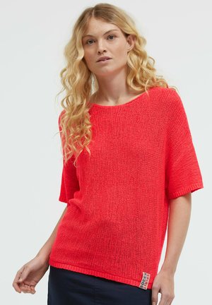 Blonde woman with long wavy hair wearing a bright red short-sleeve knit top and dark pants against a plain background.
