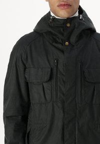 Man wearing a black hooded jacket with two zippered chest pockets and flap pockets, buttoned at the collar over a white turtleneck.