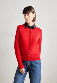 Red cardigan with a round neckline, button-down front, and ribbed cuffs, worn over a black and white striped shirt. Casual fit.