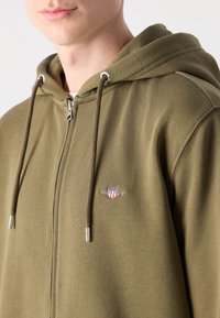 Groene rits hoodie met een logo op de borst, een capuchon met trekkoord en accenten van zilverkleurig metaal. Zachte katoenen stofmix.
