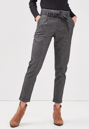 Persoon draagt een grijze broek met hoge taille en een gestrikte riem, een zwart geribbeld langärmelig shirt en bruine suède enkellaarzen.