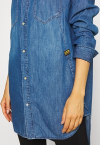 Blue denim long-sleeve shirt with snap buttons, two chest pockets, rolled-up sleeves, and a black and yellow G-Star Originals tag on the side.