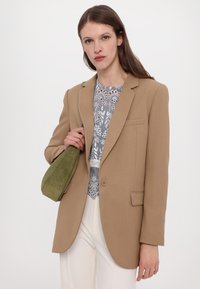 Beige räätälöity bleiseri, jossa on lovet, kuvioidun paidan päällä, valkoisten housujen ja vihreän nahevuoron olkalaukun kanssa.
