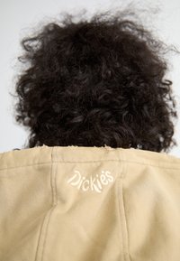 Pessoa com cabelo encaracolado escuro, a usar uma jaqueta bege com um logótipo branco bordado "Dickies" na parte de trás.