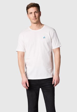 Man draagt een eenvoudig wit T-shirt met korte mouwen en een klein blauw tractorlogo op de borst, gecombineerd met zwarte jeans, staand tegen een grijze achtergrond.