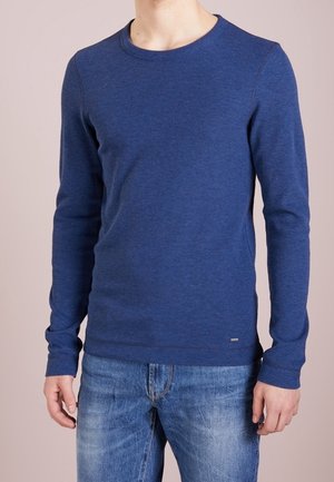 Uomo che indossa una maglia aderente a maniche lunghe blu con scollo rotondo e jeans blu, in piedi contro uno sfondo chiaro e uniforme.